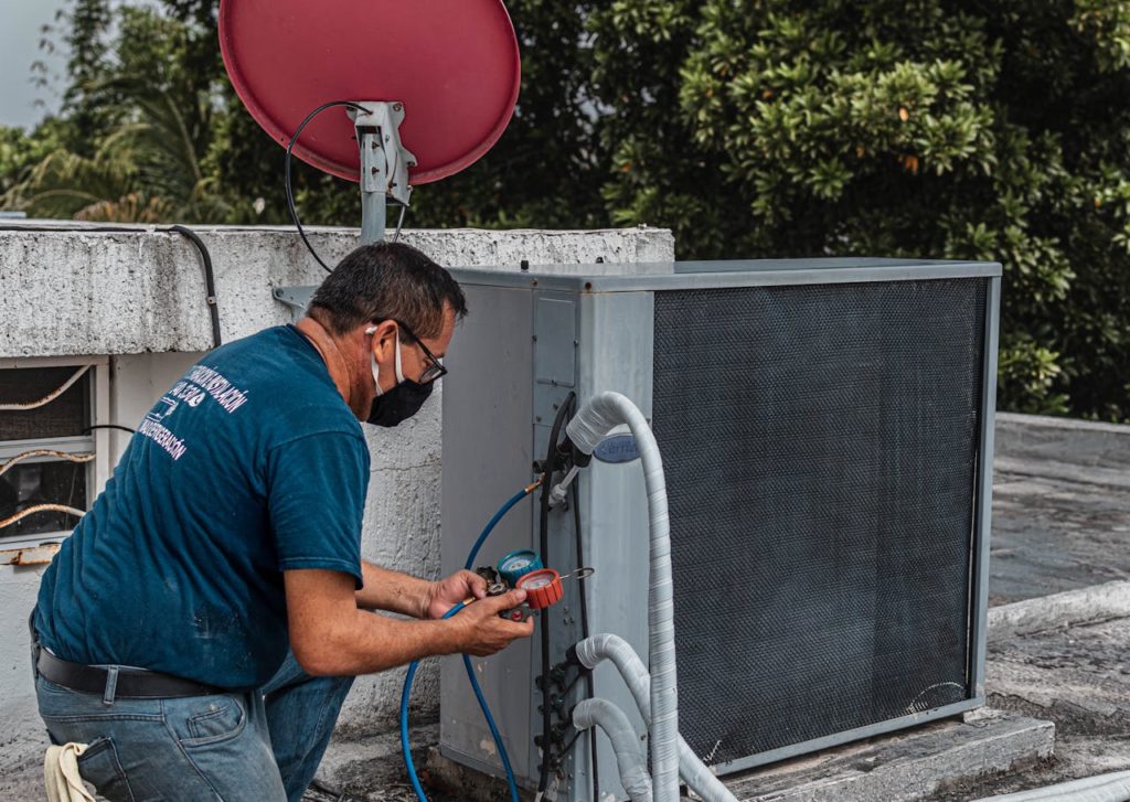 Técnico ajoelhado em um telhado, usando ferramentas para manutenção de um ar-condicionado, com mangueiras conectadas ao equipamento e uma antena parabólica ao fundo.