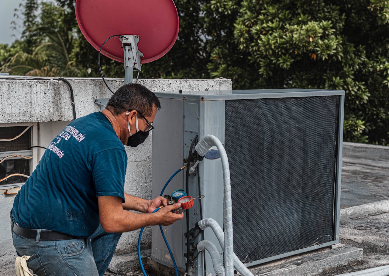 Técnico ajoelhado em um telhado, usando ferramentas para manutenção de um ar-condicionado, com mangueiras conectadas ao equipamento e uma antena parabólica ao fundo.