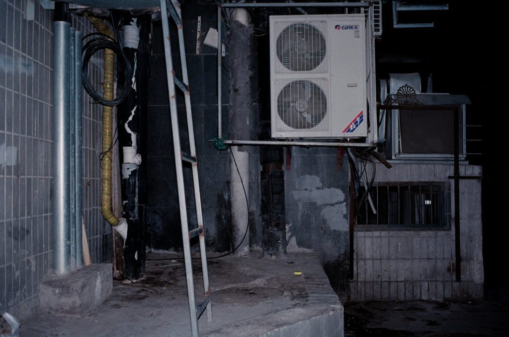 Unidade de ar-condicionado GREE montada em uma parede com suporte metálico, ao lado de uma escada e tubulações, em um ambiente industrial ou de manutenção.