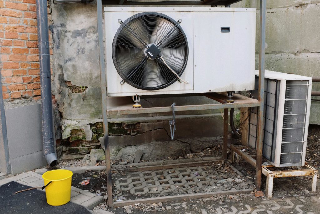 Unidade externa de ar-condicionado com ventilador metálico e balde amarelo ao lado
