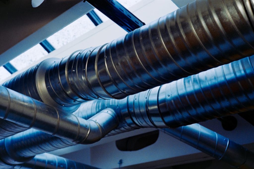 Rede de dutos metálicos de ventilação em teto industrial iluminado por luz natural.